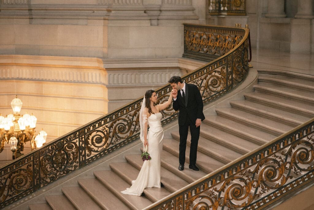 Groom kissing bride's hands on grand staircase in San Francisco City Hall