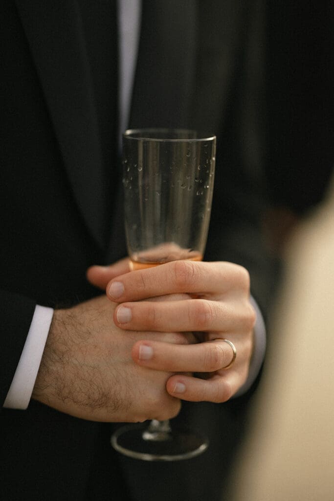 Groom holding glass of champagne during cocktail hour and showcasing ring