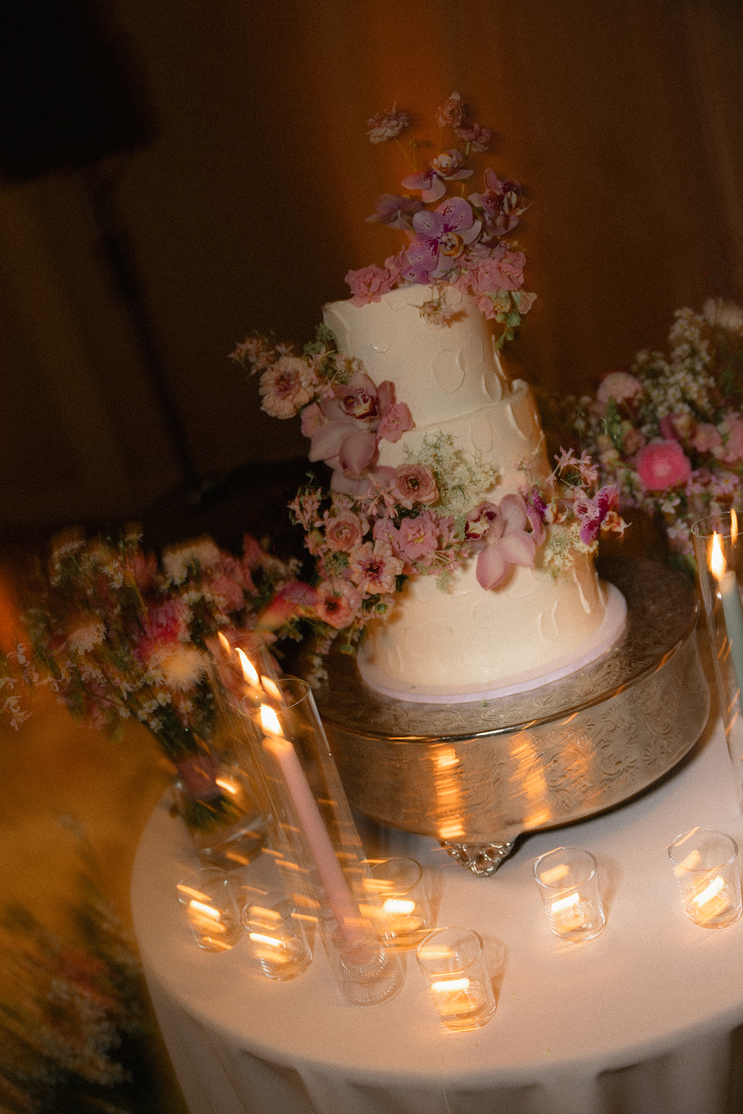Three-tier white wedding cake decorated with cascading pink orchids and fresh flowers on a candlelit reception table at casa real at ruby hill winery, photographed by a bay area wedding photographer.