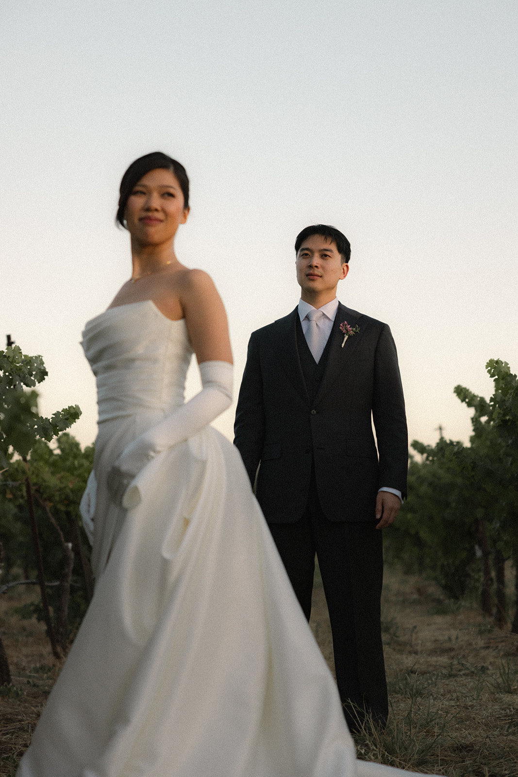 Bride in the foreground with the groom standing among vineyard rows at sunset, creating romantic casa real at ruby hill winery photos.