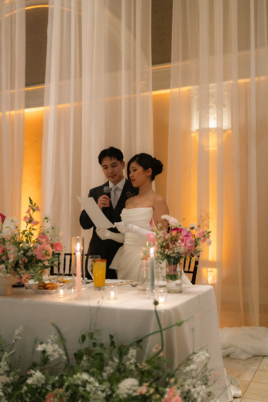 Bride and groom read their thank-you speech together at their candlelit sweetheart table surrounded by floral centerpieces at casa real at ruby hill winery, photographed by a bay area wedding photographer.