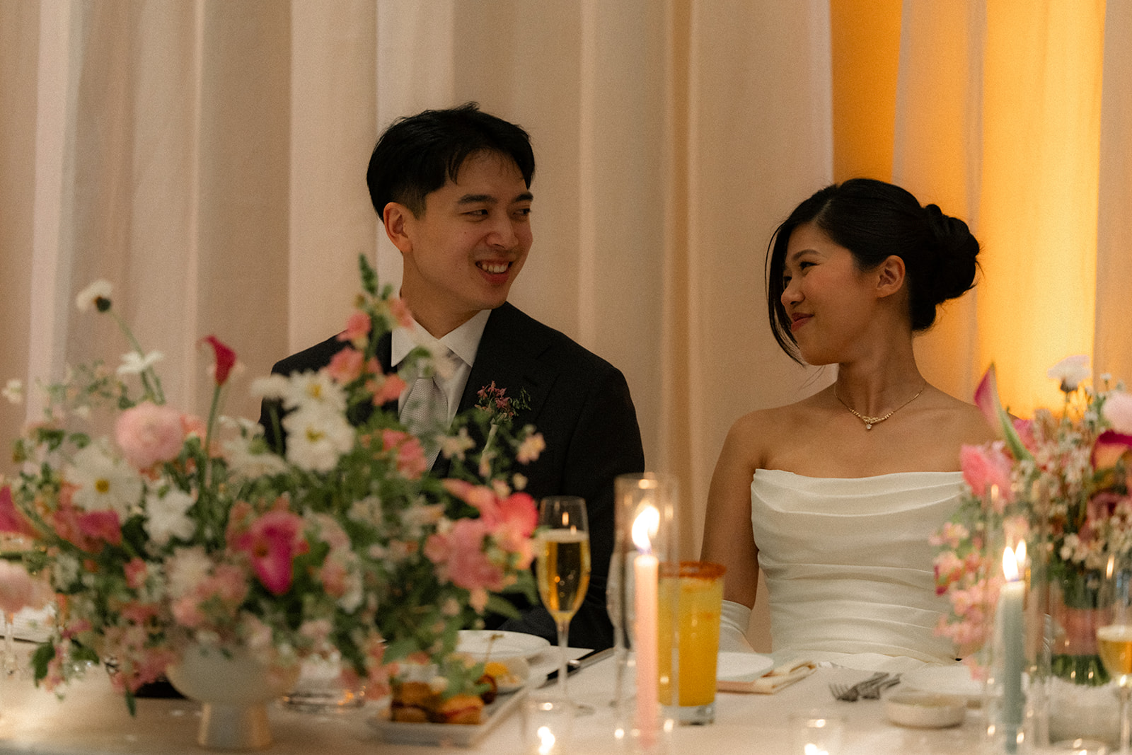 Bride and groom smile at each other during their reception dinner surrounded by pastel floral arrangements and candlelight at casa real at ruby hill winery, photographed by a bay area wedding photographer.