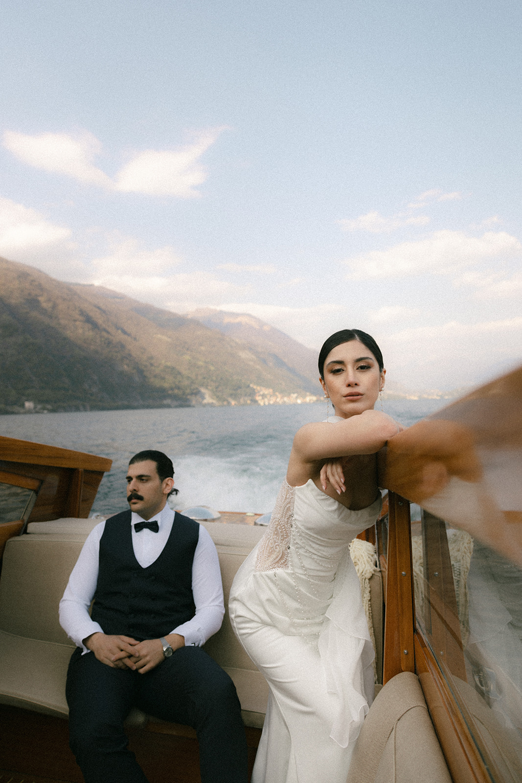 Bride posing at the side of a wooden boat while the groom sits nearby, with mountains and the lake creating a scenic wedding portrait backdrop.