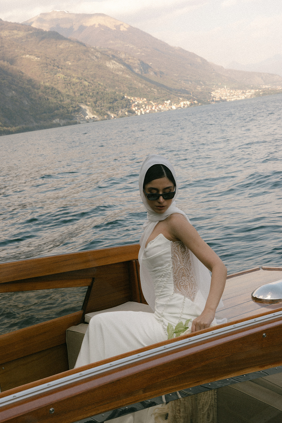 Bride wearing a silk headscarf and sunglasses aboard a vintage wooden boat, photographed during an Italian destination wedding on the lake.