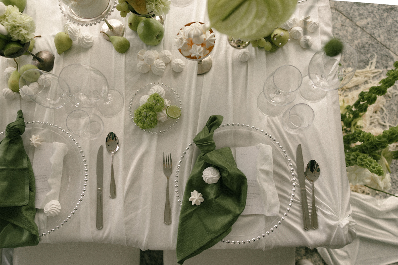 Overhead view of an elegant wedding reception table featuring white linens, green napkins, glassware, fresh fruit, and decorative desserts.