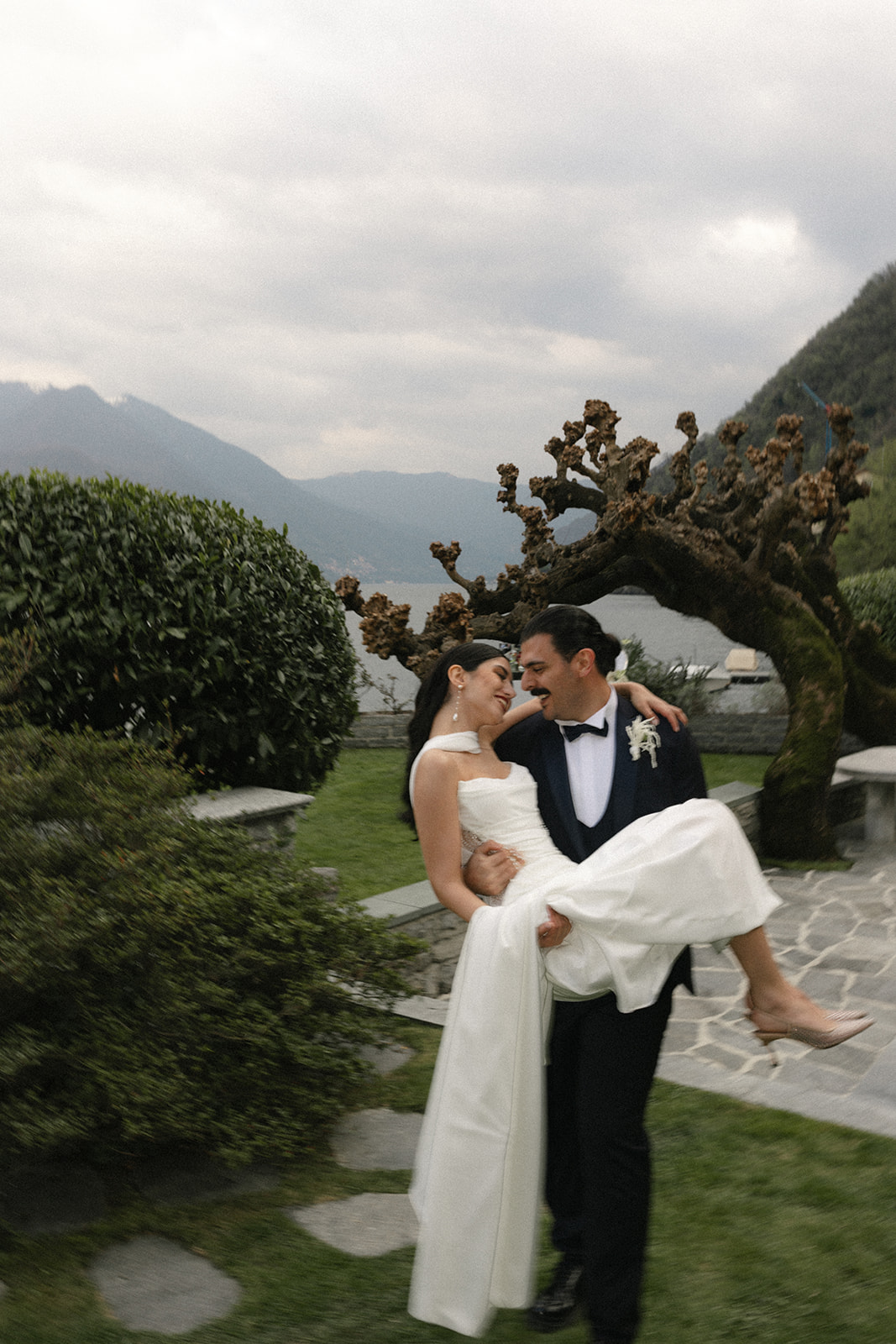 Groom carrying the bride through a lakeside garden during an Italian destination wedding, captured by a destination wedding photographer with Lake Como in the background.