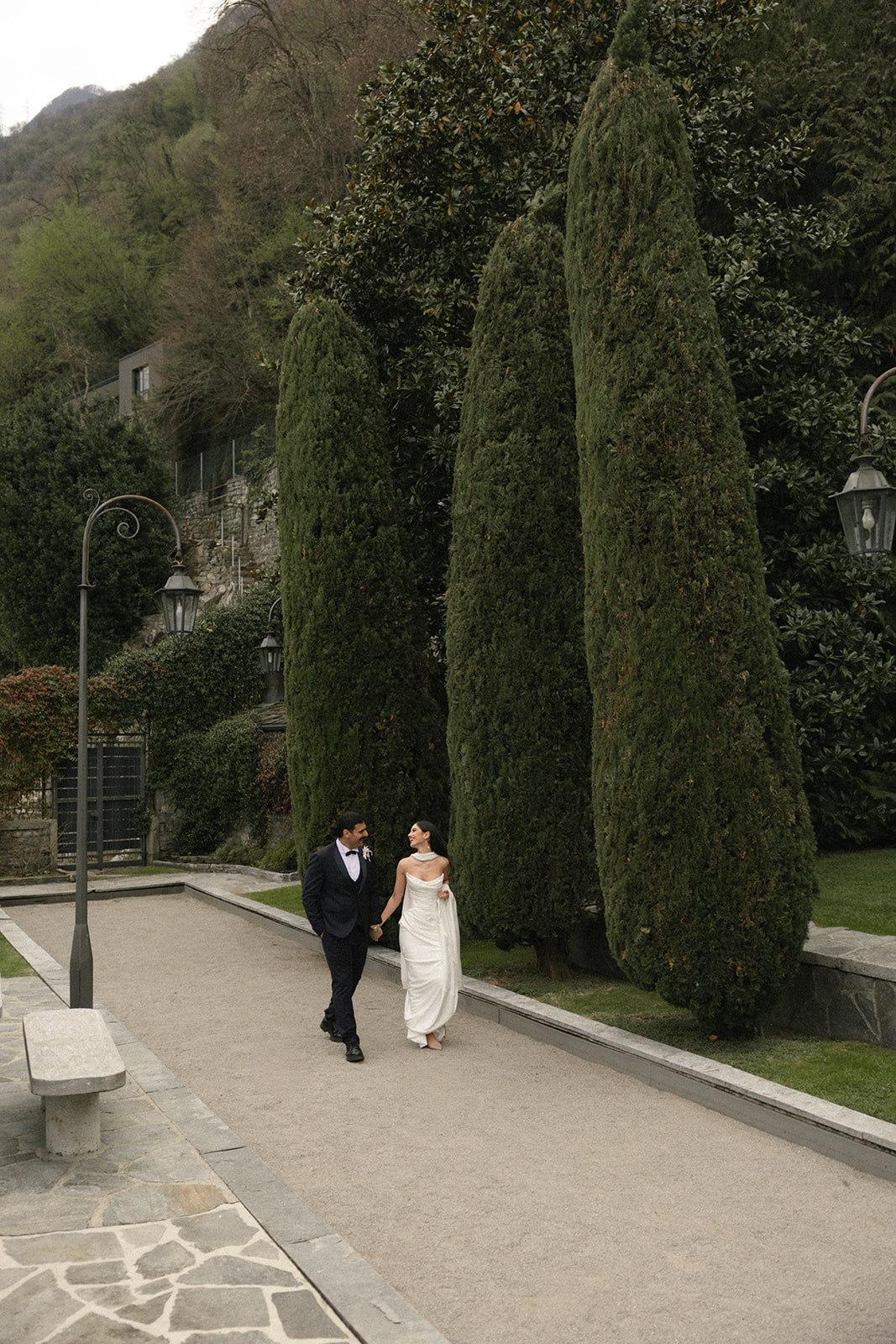 Bride and groom walking hand in hand through the gardens during a Lake Como wedding, surrounded by tall cypress trees.