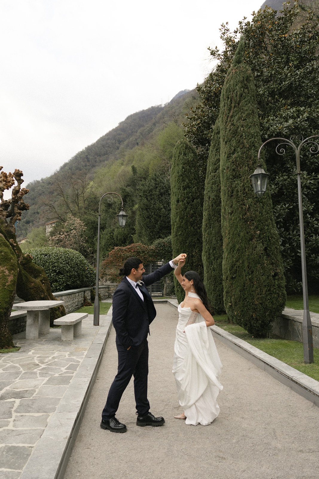 Bride twirling beneath the groom’s raised hand along a garden pathway lined with tall cypress trees during a Lake Como wedding.