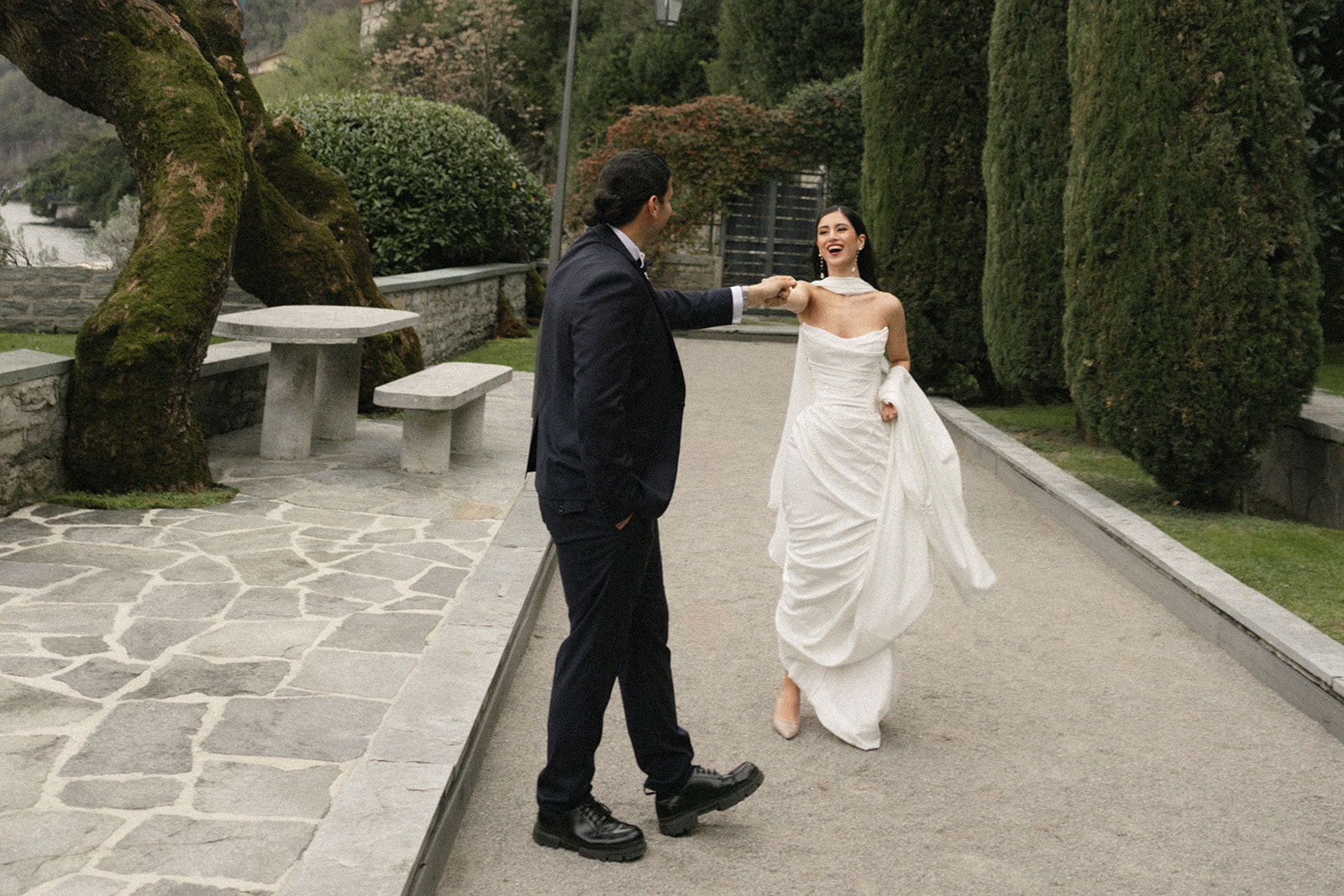 Bride smiling as the groom gently twirls her along a garden pathway lined with cypress trees, creating a playful and romantic wedding portrait.