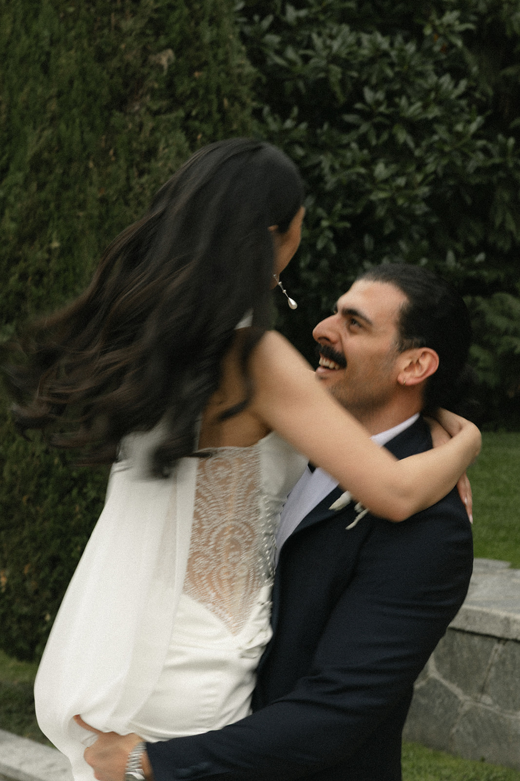 Groom lifts the bride into an embrace as they smile at each other in a candid garden wedding portrait surrounded by lush greenery.