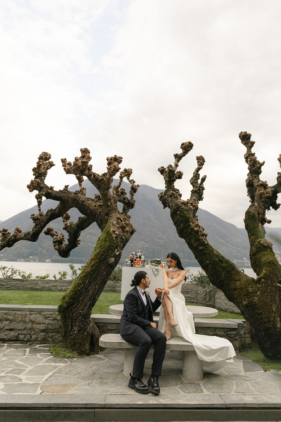 Bride and groom seated at a stone picnic table beneath sculptural moss-covered trees, holding hands during an intimate garden wedding portrait.
