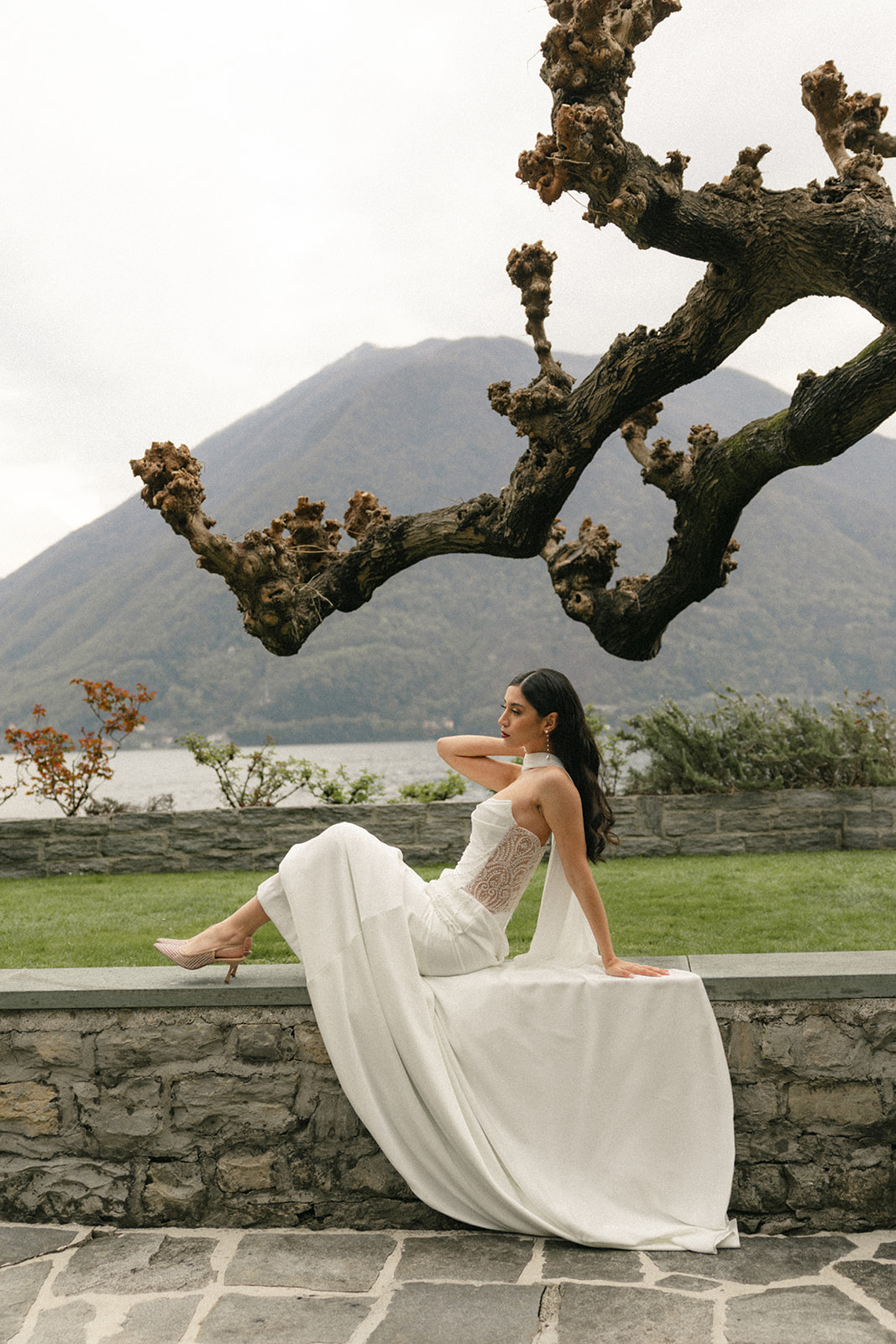 Bride seated on a stone garden wall beneath a sculptural tree, with mountain and lakeside views creating an elegant wedding portrait.