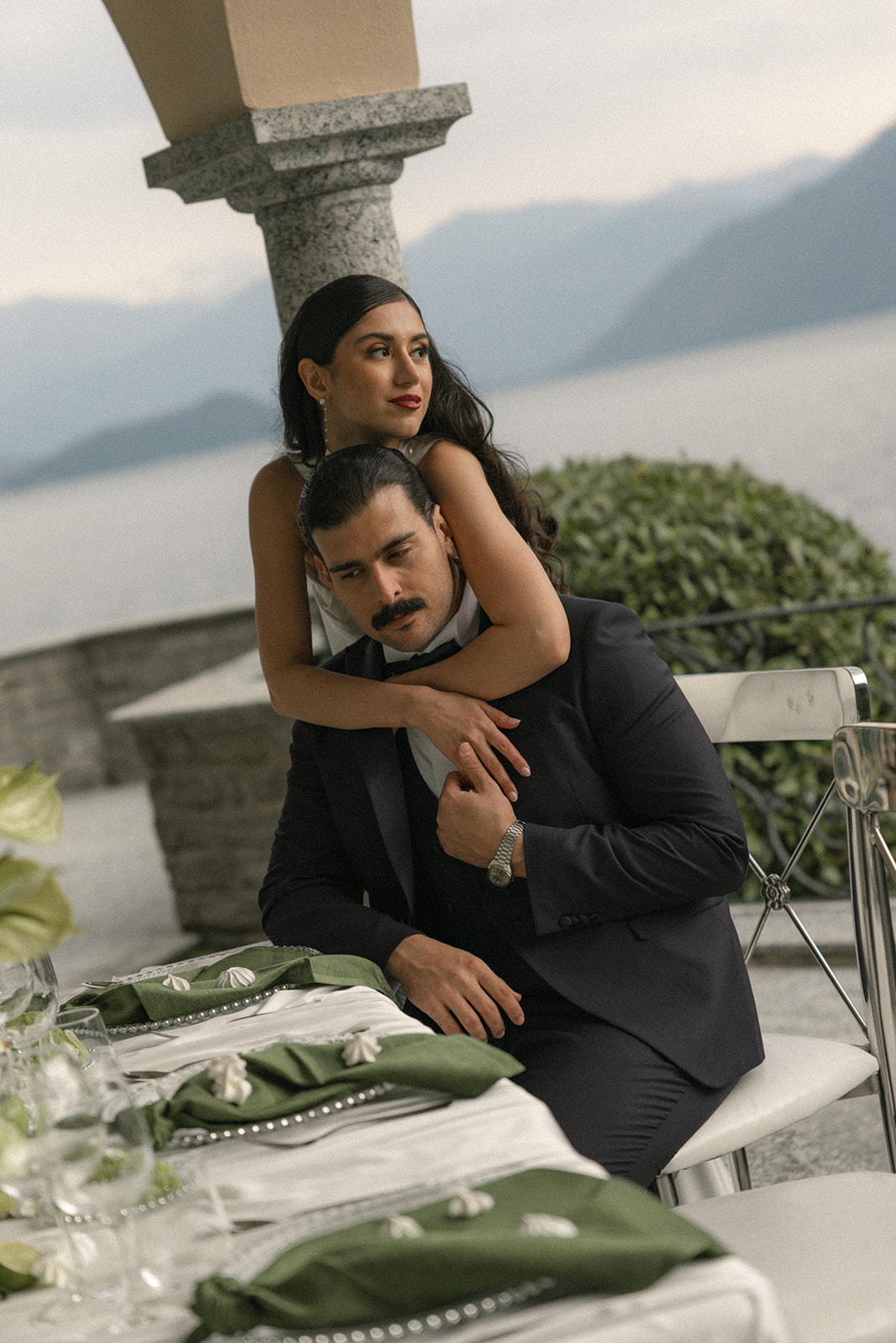 Bride embracing the seated groom at an elegant outdoor wedding reception table overlooking a lake and mountains.