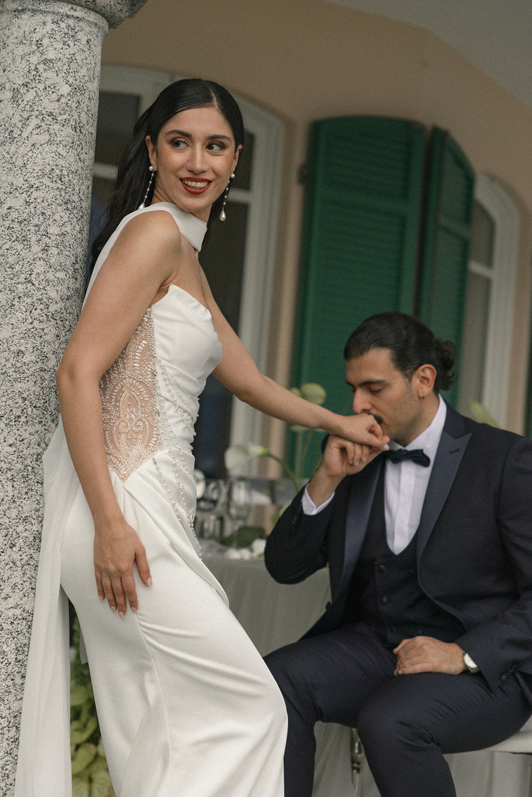Bride standing beside a stone column as the seated groom kisses her hand during an elegant outdoor wedding portrait.