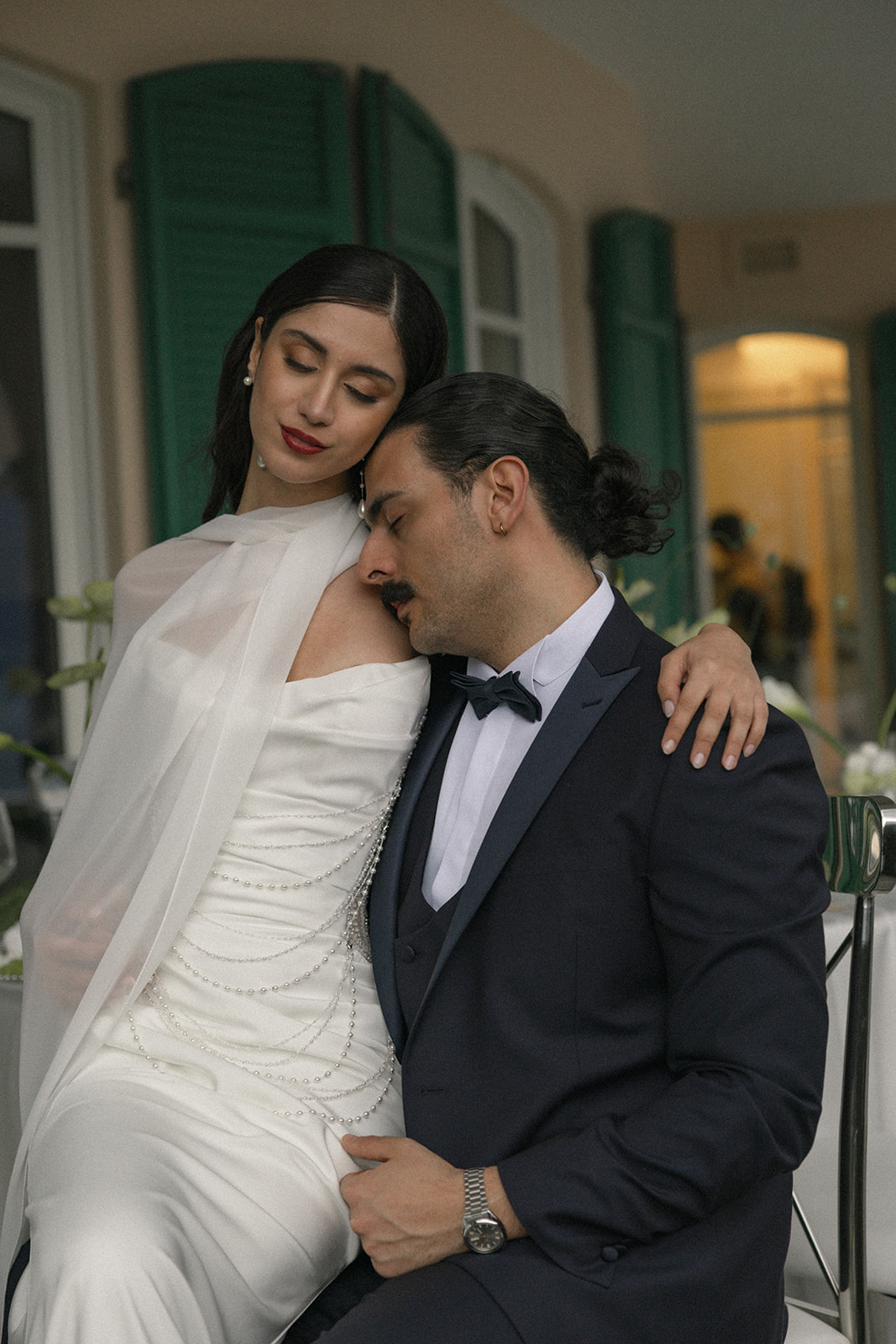 Bride and groom sharing a quiet embrace while seated together at a wedding reception, with elegant décor and villa architecture in the background.