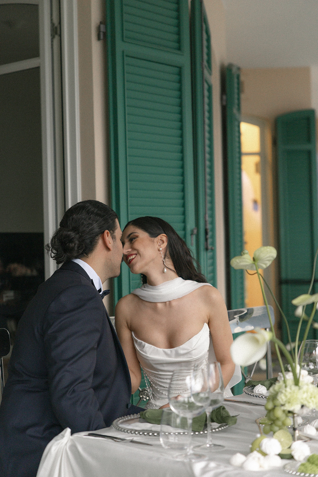 Bride and groom leaning in for a kiss while seated at an elegantly styled reception table with green linens, white florals, and glassware.
