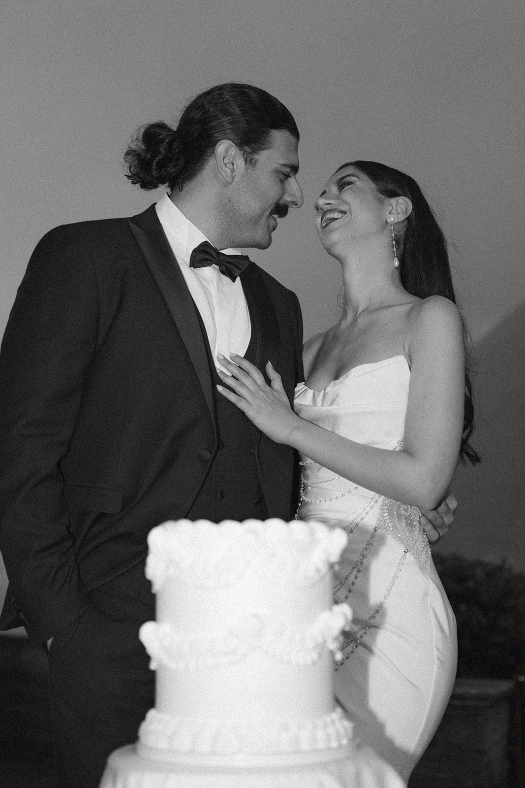 Bride and groom smiling at each other beside a tiered wedding cake during an evening wedding celebration.