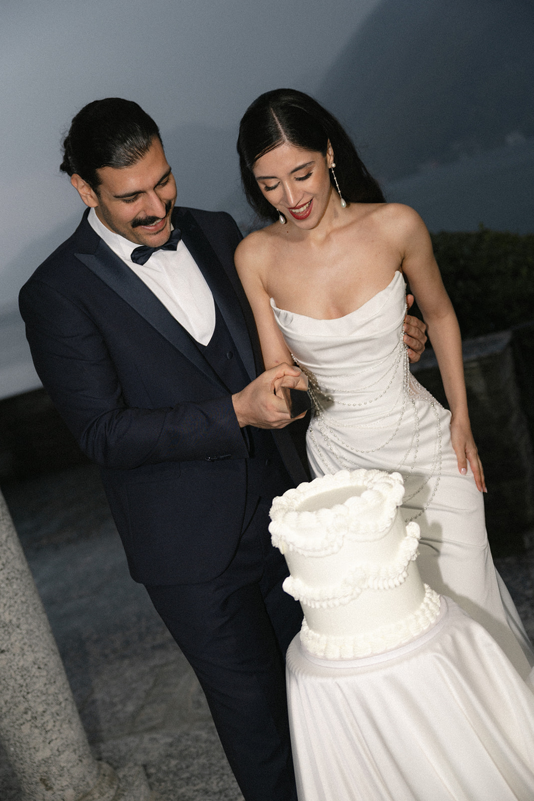 Bride and groom smiling together as they cut a white tiered wedding cake during an evening outdoor wedding celebration.