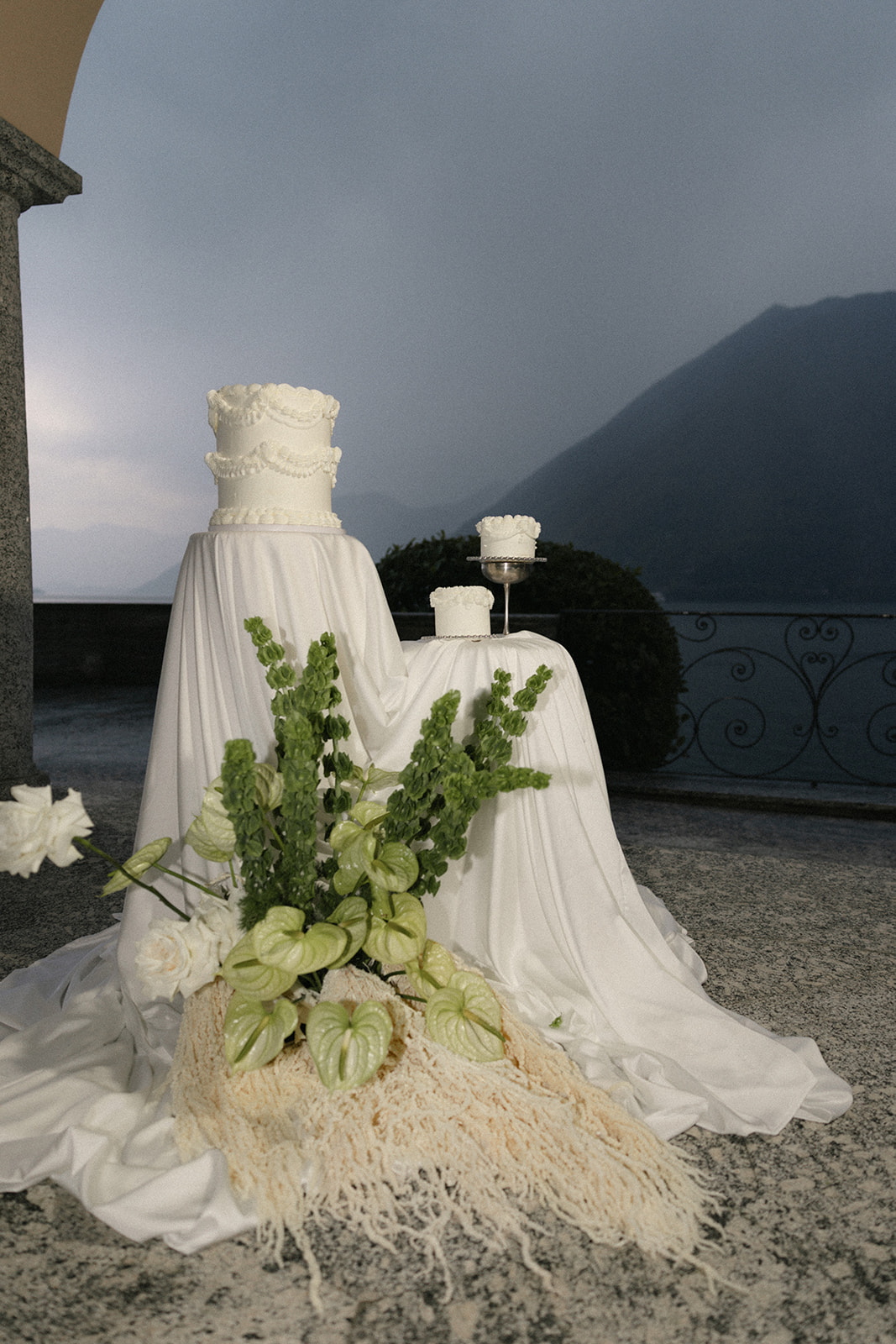 Elegant white wedding cake display with fresh green and white floral arrangements overlooking a lake and mountain landscape at dusk.
