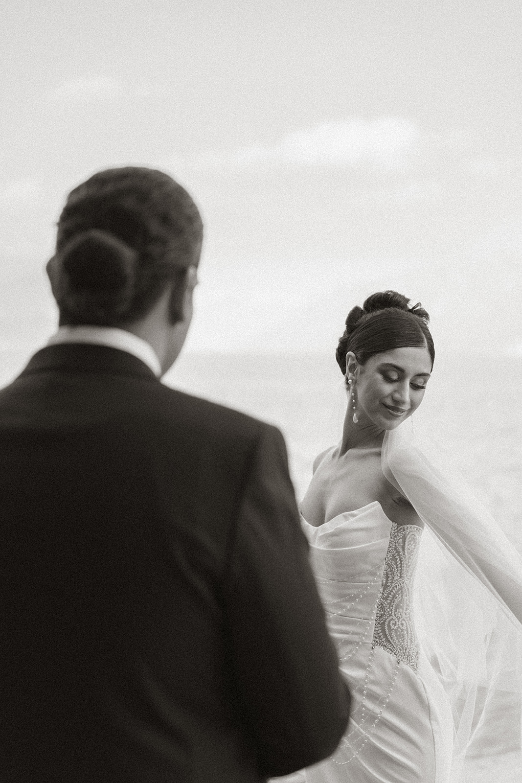 Bride smiling over her shoulder as her veil flows in the breeze, photographed in a timeless black-and-white portrait by a destination wedding photographer.