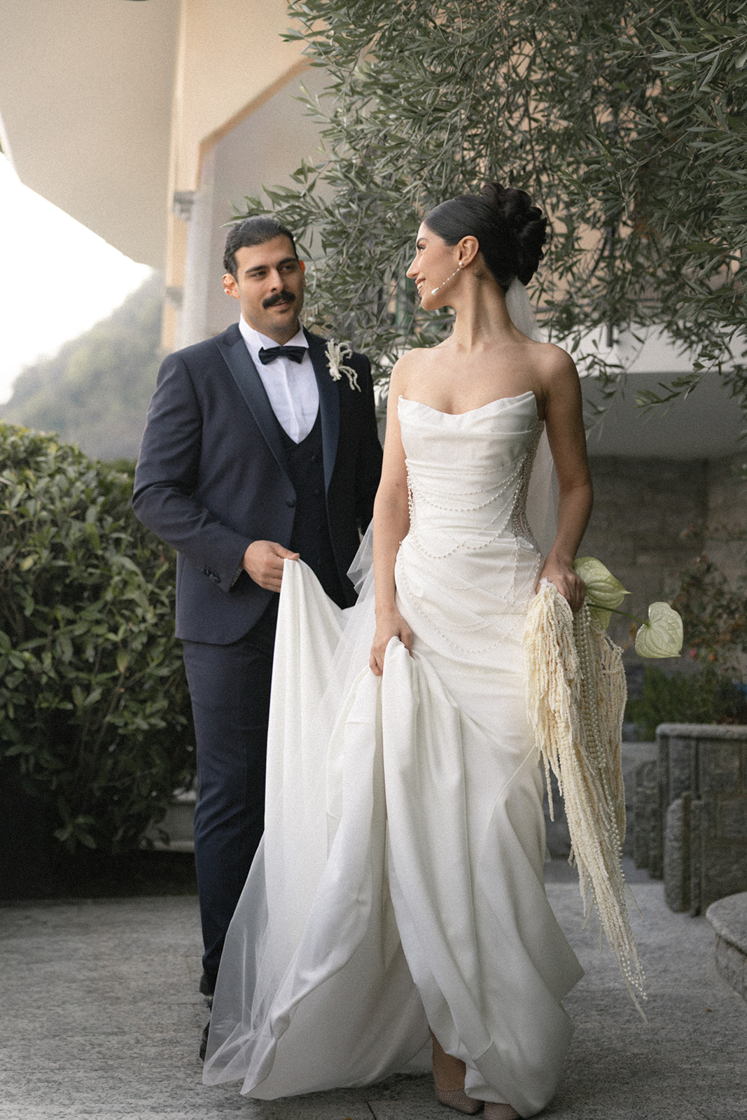Bride walking ahead while the groom carries the train of her gown during a Lake Como wedding, surrounded by lush gardens and elegant floral details.