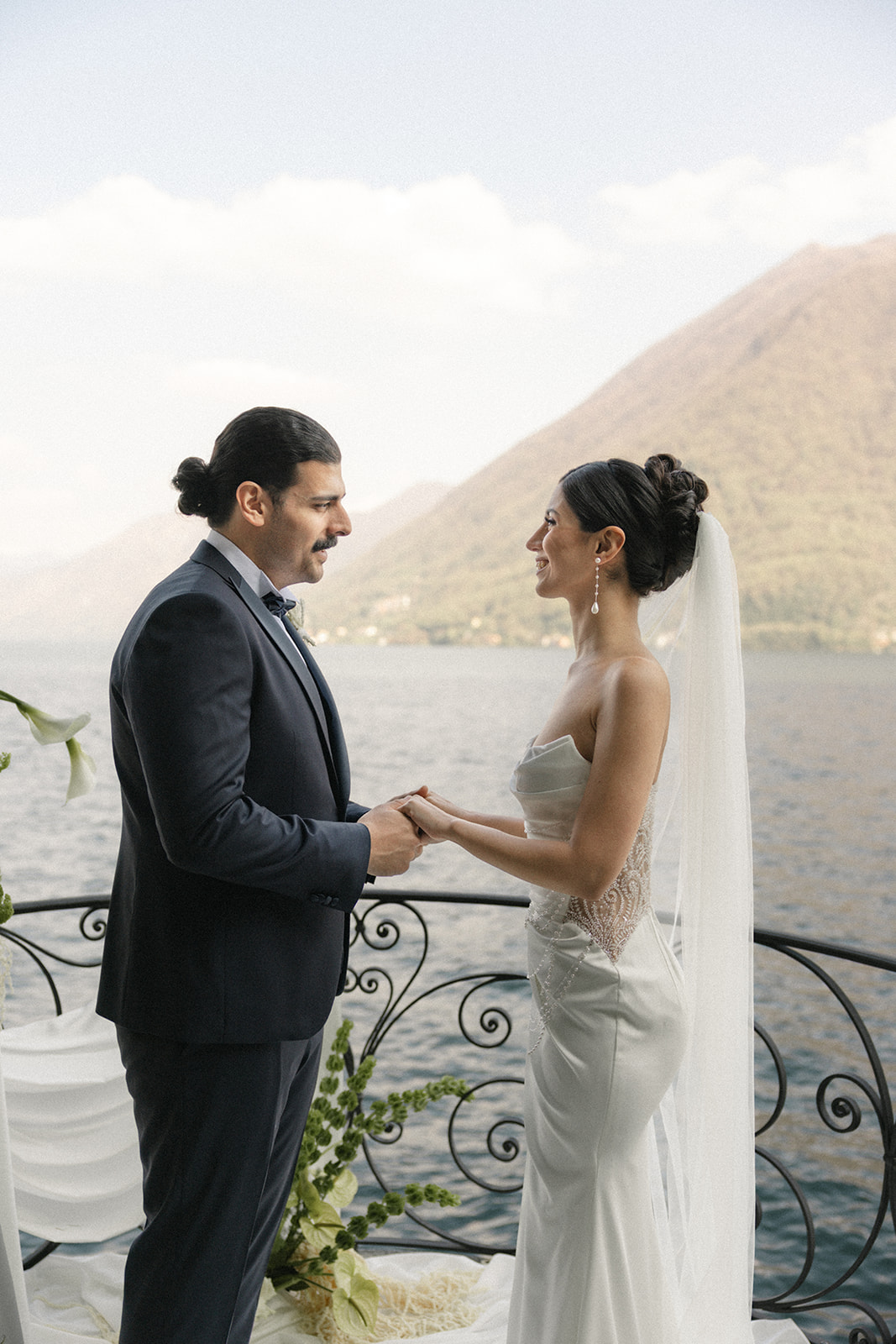 Bride and groom holding hands on a lakeside terrace with mountain views, photographed by a destination wedding photographer.