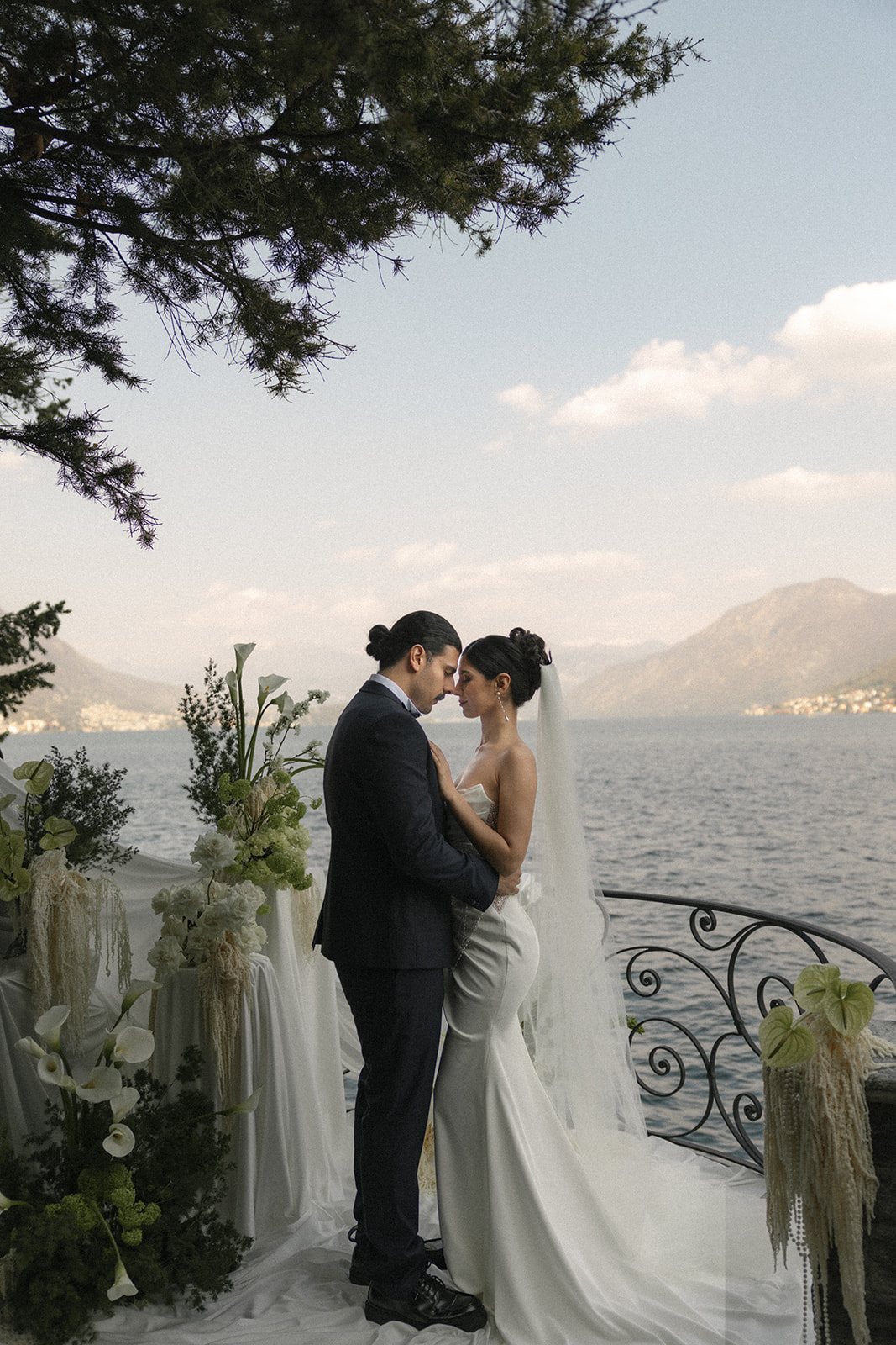 Bride and groom embracing on a lakeside terrace with floral arrangements and mountain views during an Italian destination wedding.