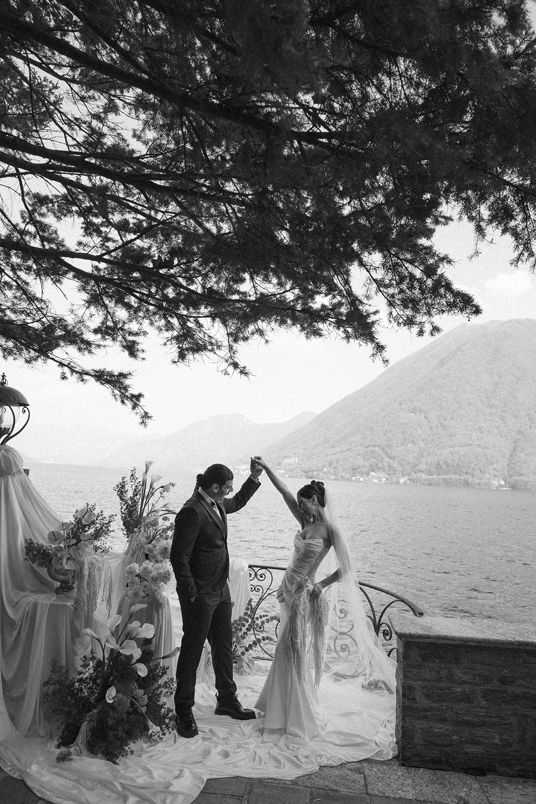 Bride twirling beneath the groom's raised hand on a flower-filled terrace overlooking the lake, photographed in a timeless black-and-white style.