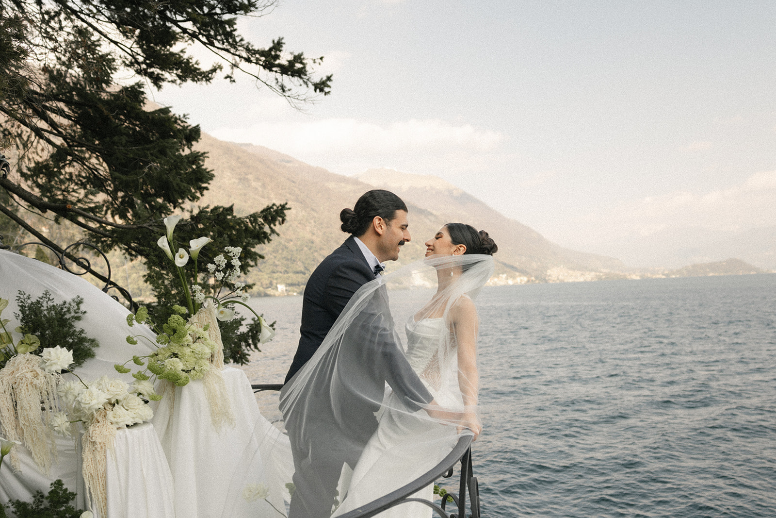 Bride and groom sharing a quiet moment on a lakeside terrace with flowing veil, overlooking the water during an Italian destination wedding.