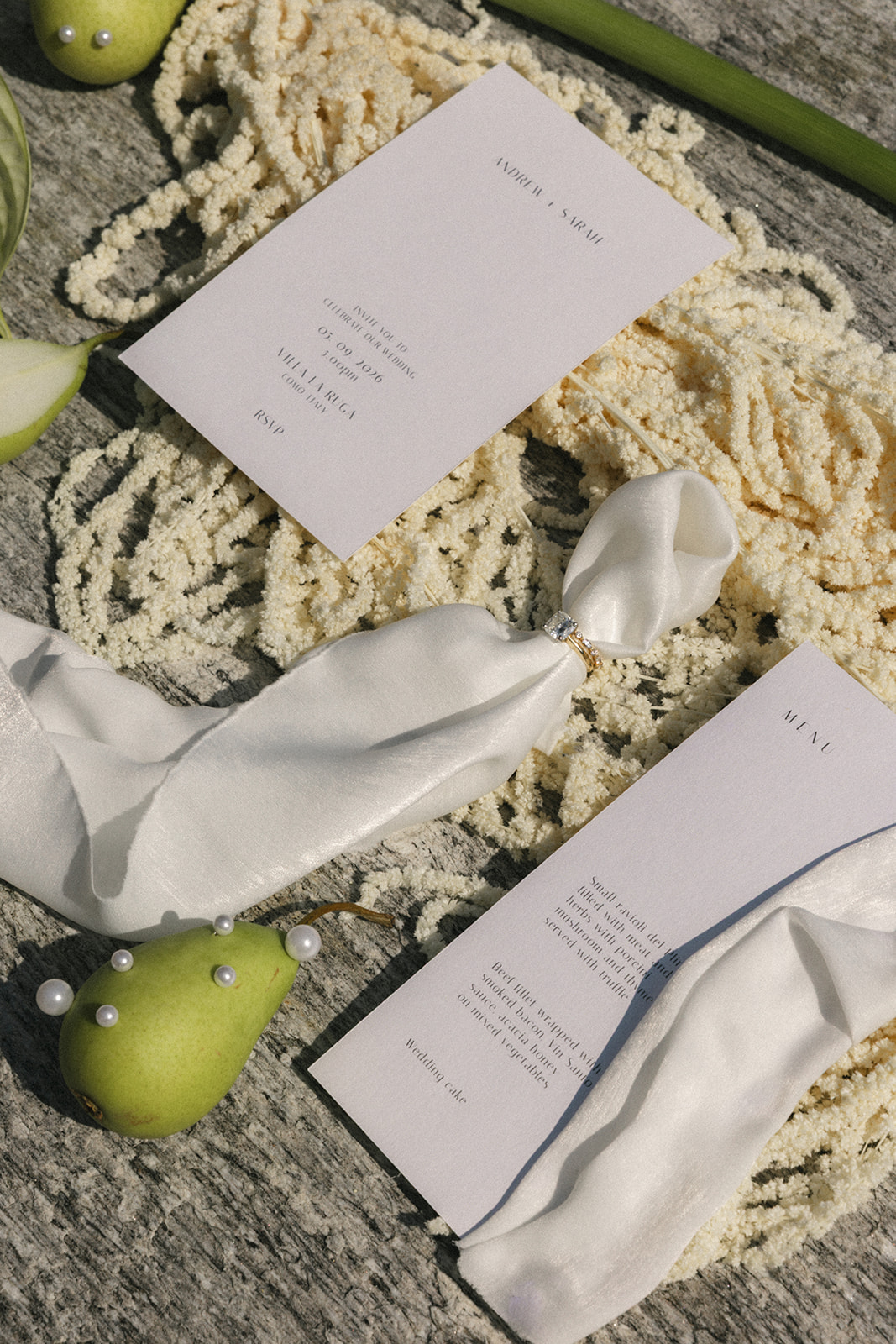 Flat lay of wedding invitation, menu card, silk ribbon, ring, pears, and floral details styled for an Italian destination wedding.
