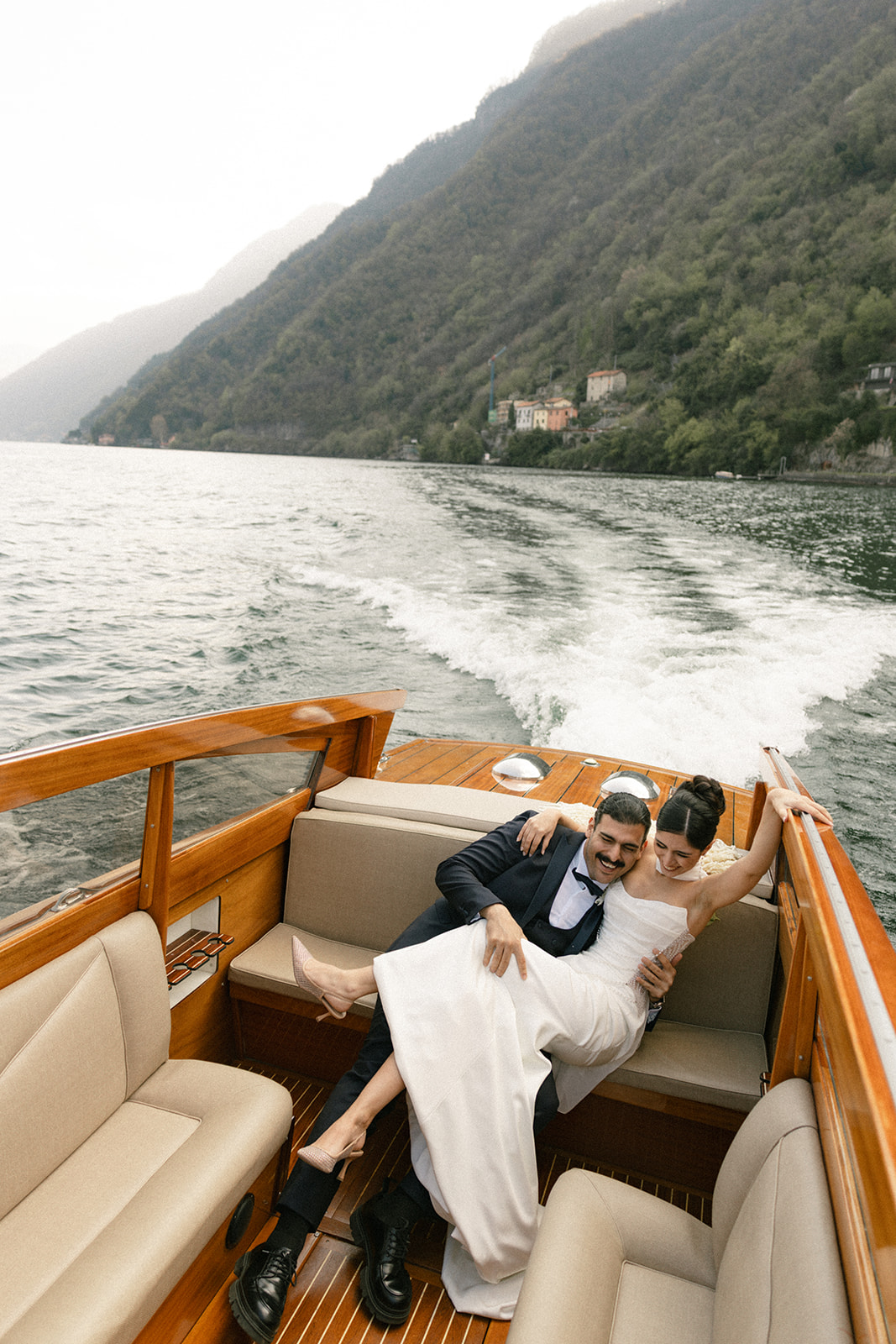 Bride and groom laughing together aboard a classic wooden boat during a Lake Como wedding, with the lake and mountains stretching behind them.
