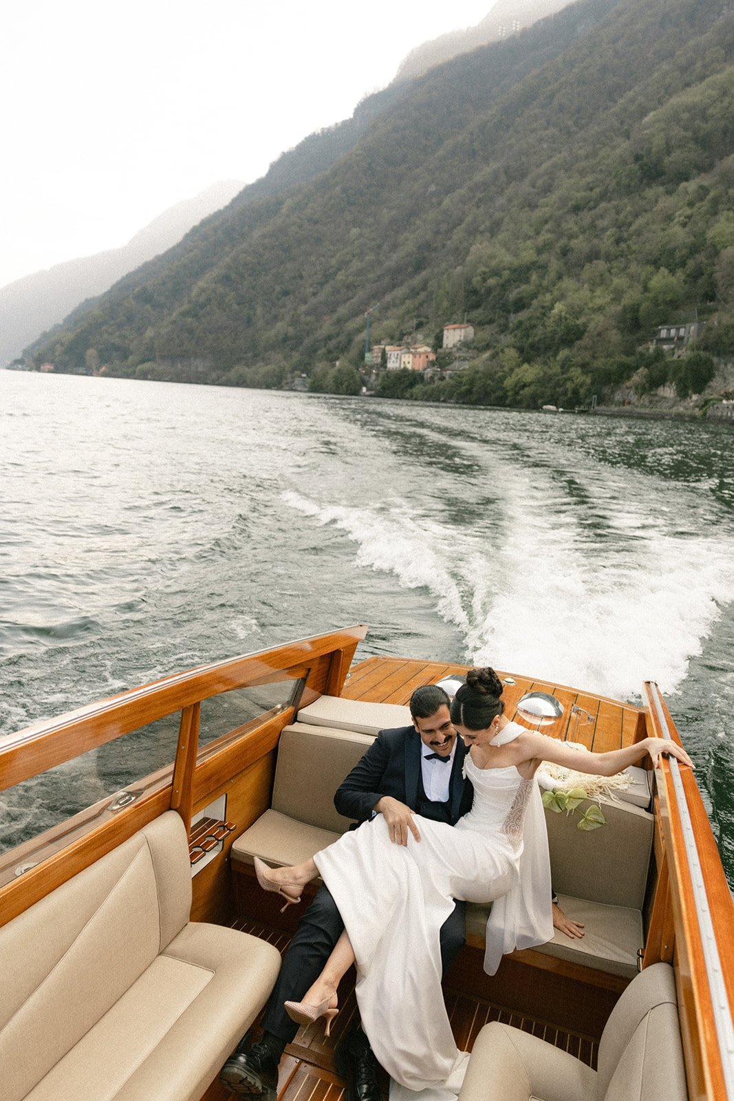 Bride and groom relaxing in a classic wooden boat on the water during a Lake Como wedding, with mountain views and the boat's wake stretching across the lake.