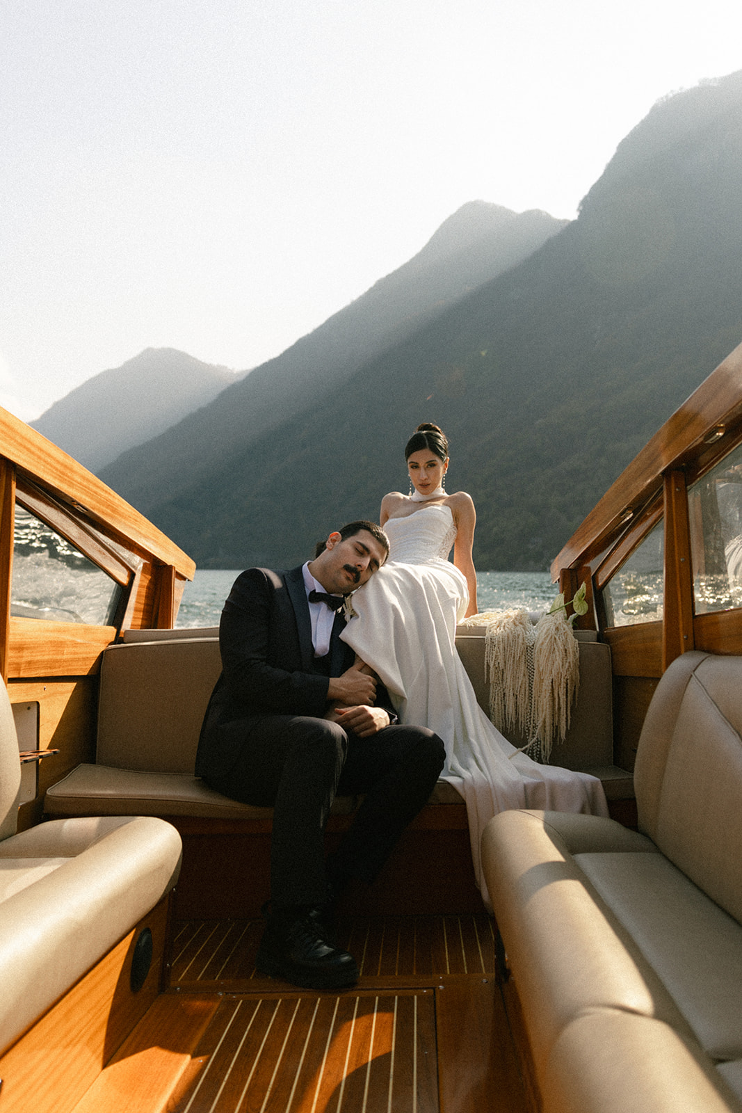 Bride and groom relaxing aboard a wooden boat, framed by mountain and lake views, during a romantic wedding portrait session.