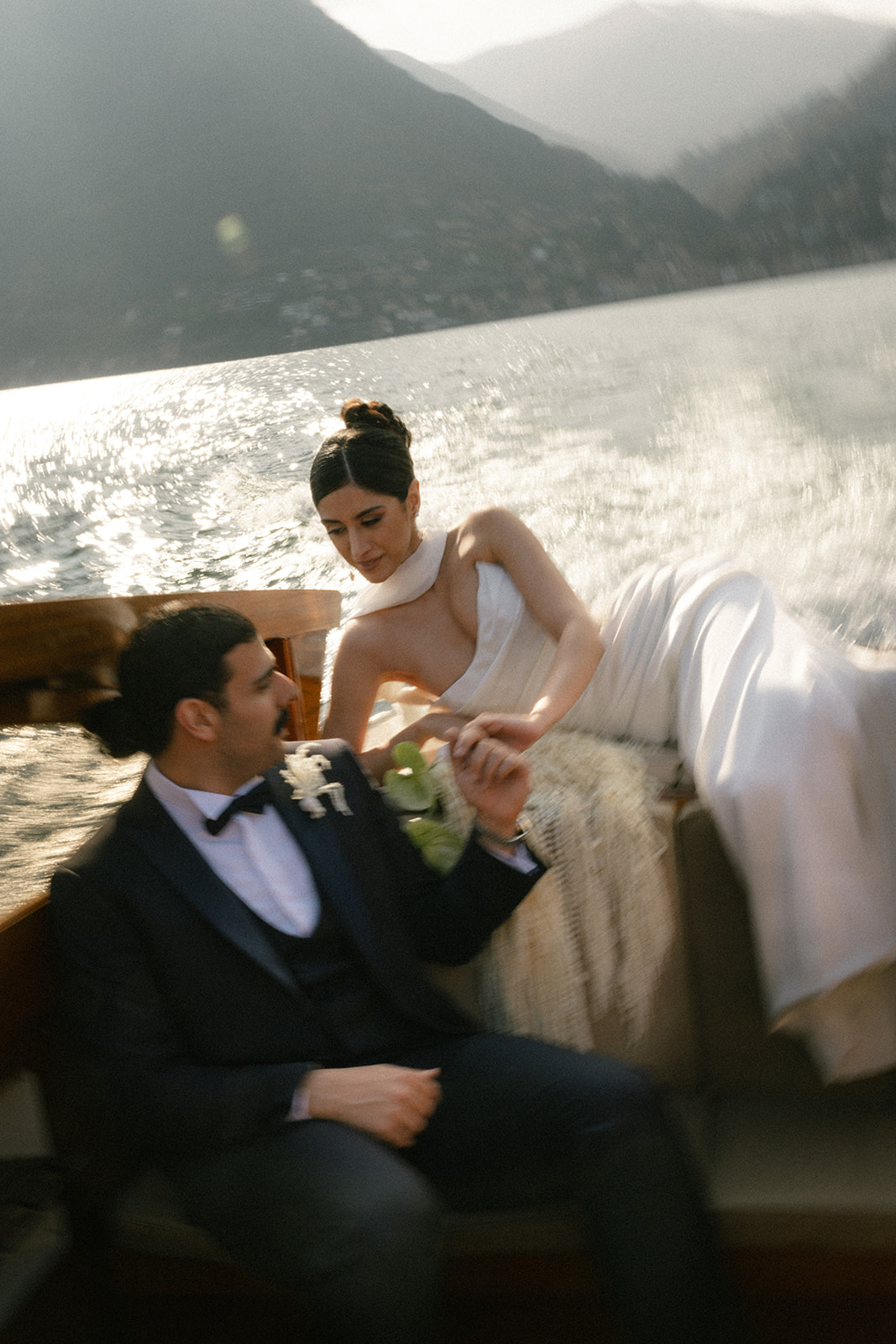 Bride and groom holding hands aboard a speeding wooden boat, with sunlight reflecting across the water during a Lake Como wedding.