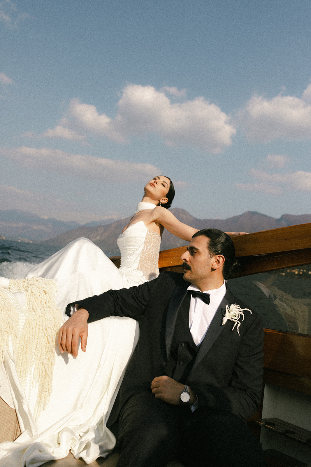 Bride and groom relaxing aboard a wooden boat with mountain views, celebrating an elegant Italian destination wedding on the lake.