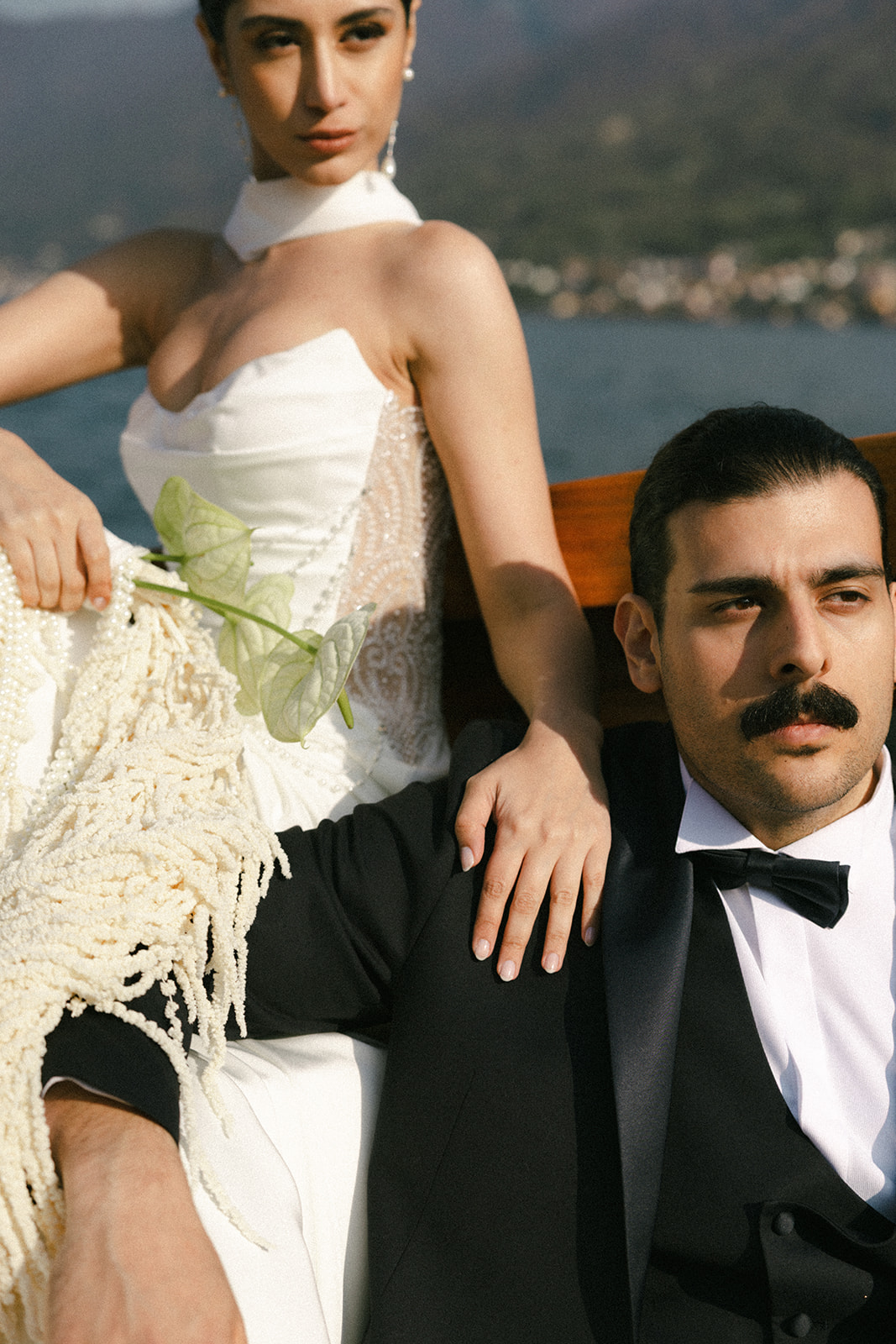 Close-up of the bride and groom seated on a wooden boat, with the bride resting her hand on the groom's shoulder against a lakeside backdrop.