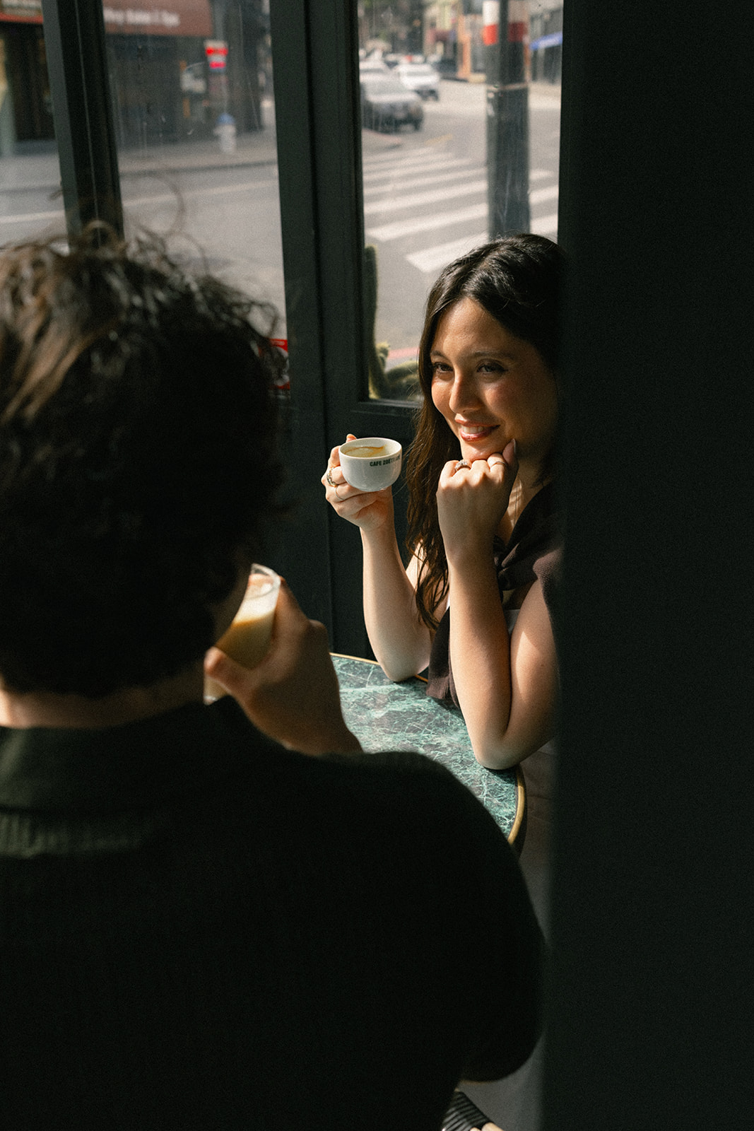 Couple sharing coffee and smiling across a café table by the window, with soft afternoon light and San Francisco City Streets visible outside during a relaxed San Francisco engagement photos session.