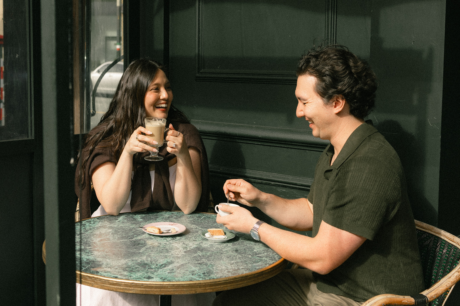 Couple laughing together over coffee at an outdoor café table, with dark storefront walls and soft afternoon light creating an intimate setting for San Francisco engagement photos.