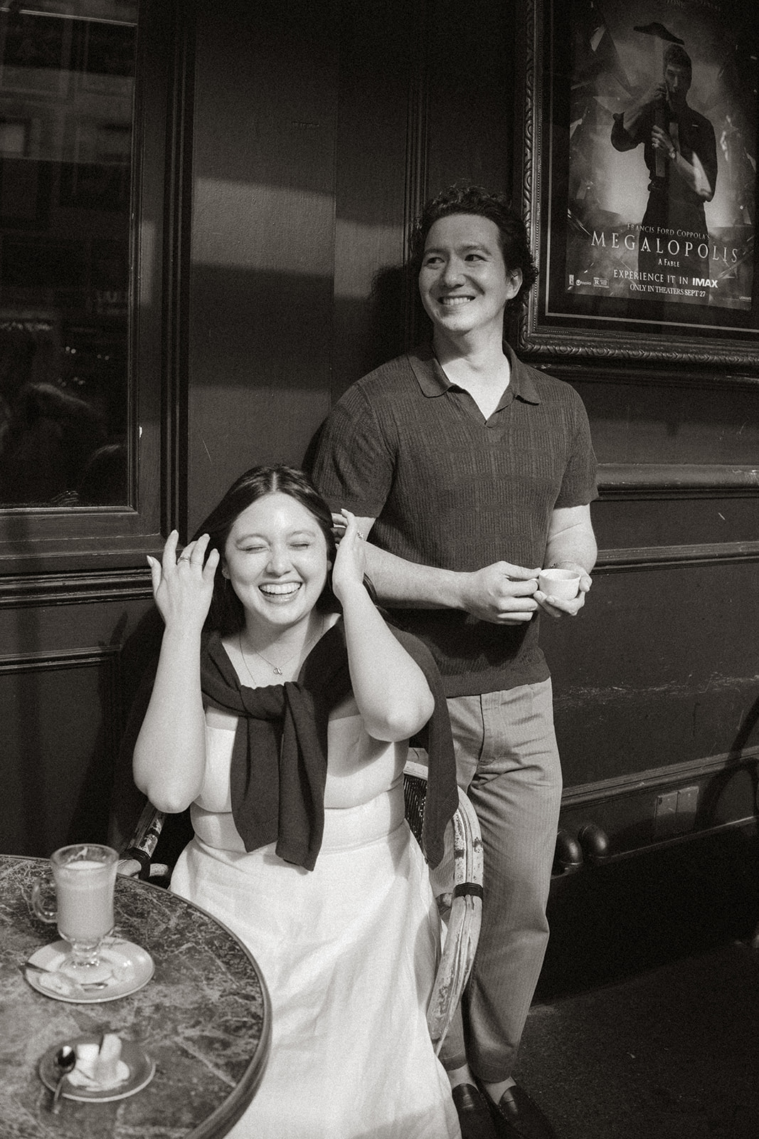 Black and white portrait of a smiling couple outside a café, with vintage movie posters and dark storefront details creating a timeless backdrop during a San Francisco engagement photos session.