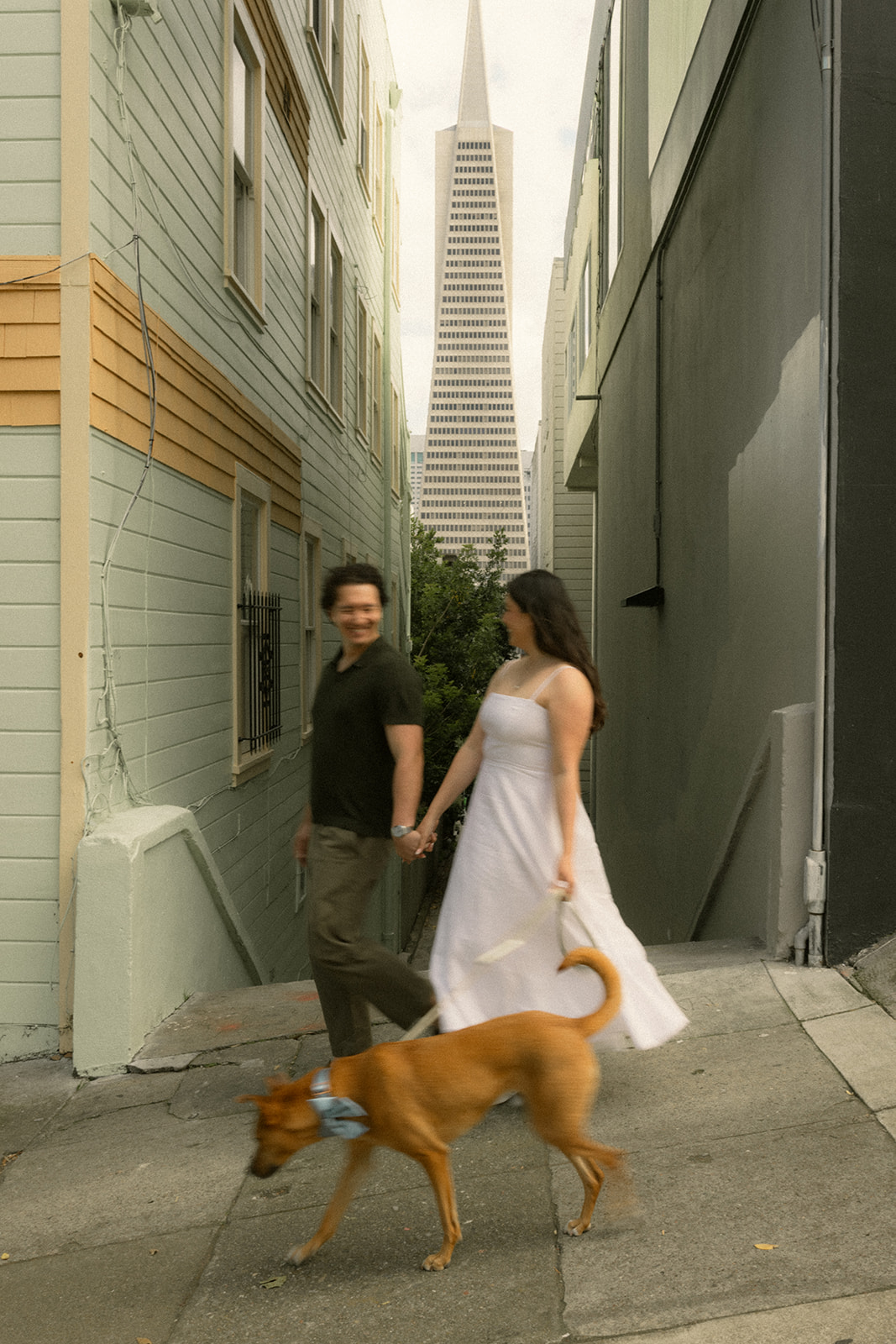 Couple walking their dog through a narrow alley framed by colorful buildings, with the Transamerica Pyramid rising in the background, photographed by a San Francisco photographer.