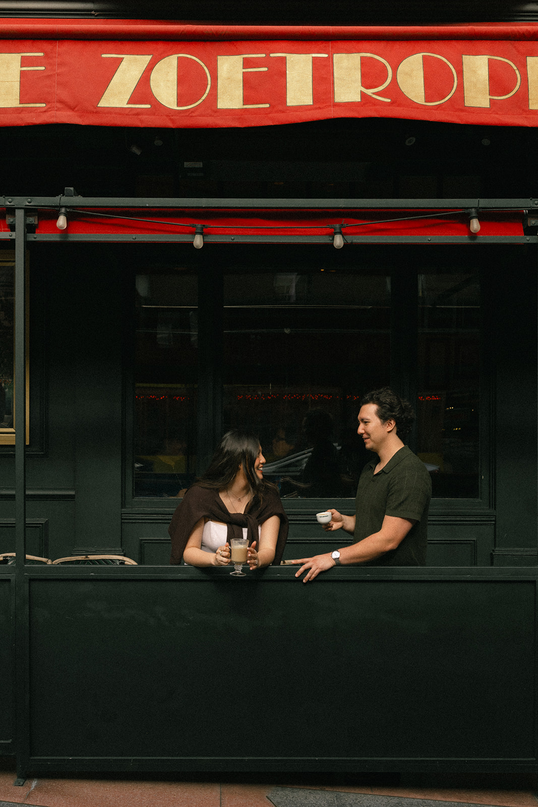 Couple enjoying coffee together outside a café beneath a red awning, smiling and talking across the patio railing photographed by San Francisco photographer