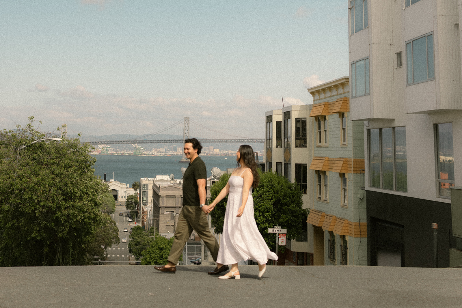 Couple holding hands while walking across a steep hill with the Bay Bridge, waterfront, and San Francisco City Streets stretching into the distance behind them during a San Francisco engagement photos session.