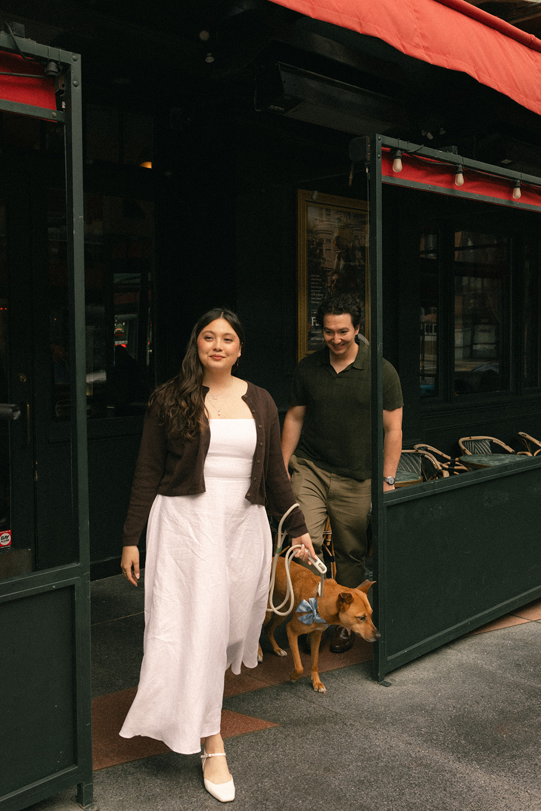 Couple leaving a downtown café with their dog, framed by a dark storefront, outdoor seating, and classic San Francisco City Streets, photographed by a San Francisco photographer.