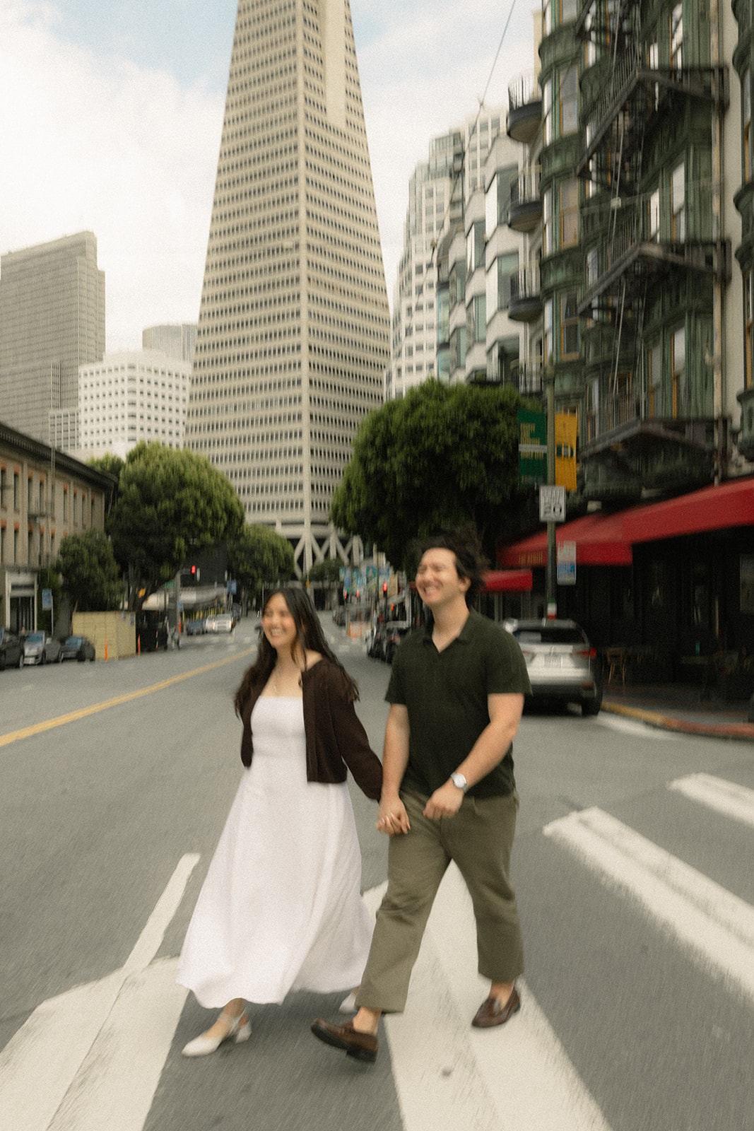 Couple walking hand in hand across a downtown street with the Transamerica Pyramid and surrounding San Francisco City Streets creating an iconic city backdrop.