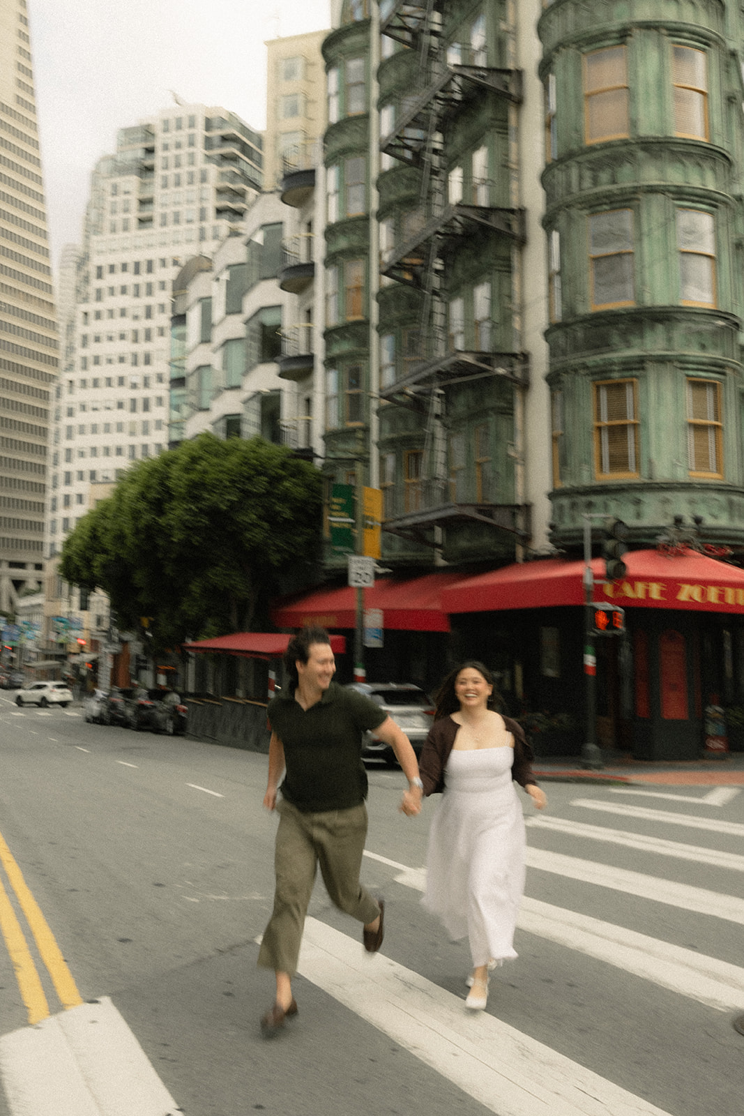 Couple running hand in hand across a downtown crosswalk beside a historic corner building in San Francisco for their downtown engagement session