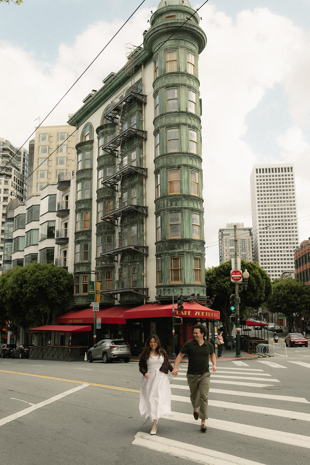 Couple holding hands while crossing the street in front of Café Zoetrope, with historic architecture and San Francisco City Streets creating an iconic downtown backdrop.
