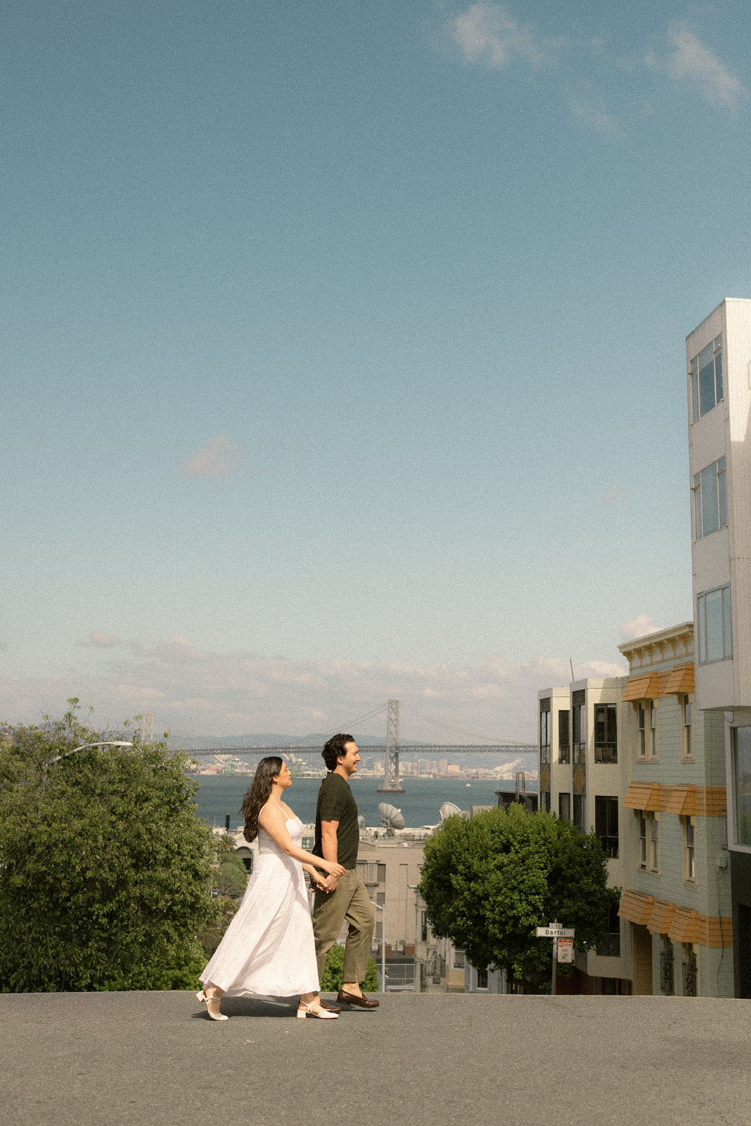 Couple walking hand in hand along a steep hill with the Bay Bridge, waterfront, and San Francisco City Streets stretching behind them during a sunny San Francisco engagement photos session.