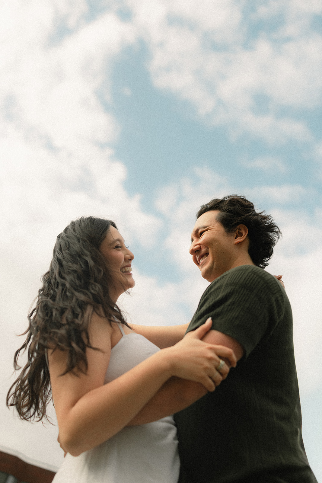 Low-angle portrait of a smiling couple embracing beneath a bright blue sky with soft clouds, creating a timeless moment captured by a San Francisco photographer.