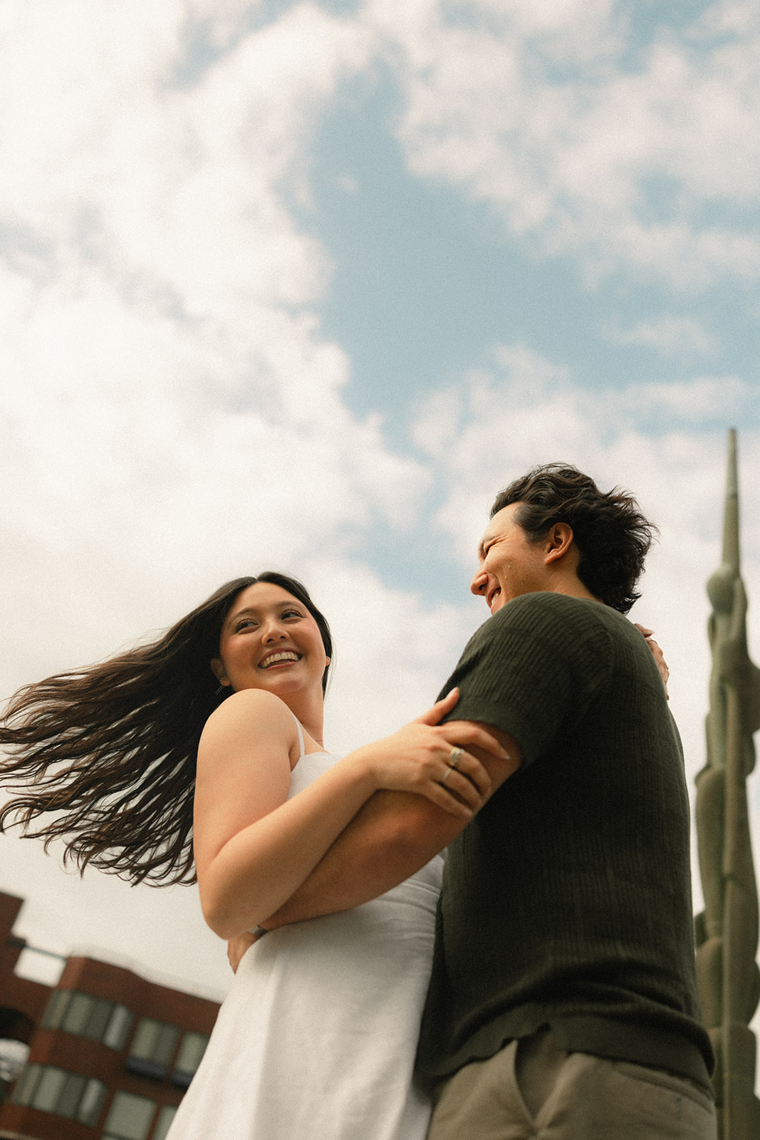 Low-angle portrait of a couple embracing beneath a bright sky, with modern city architecture softly visible in the background, captured by a San Francisco photographer.