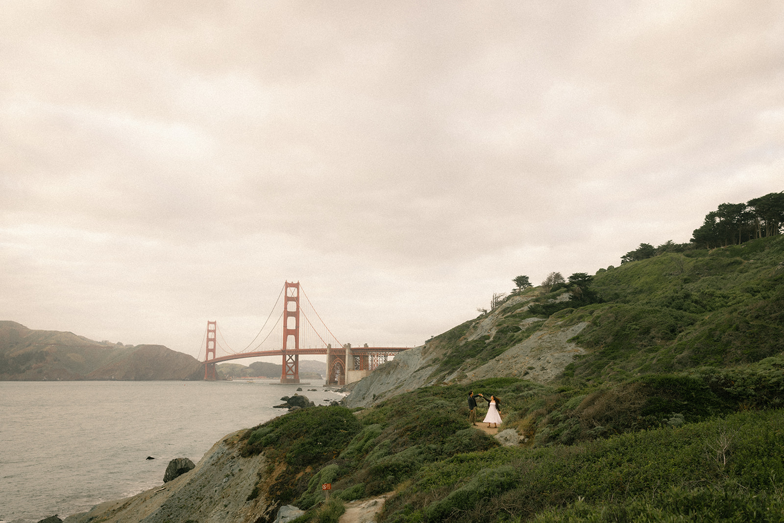 Wide landscape view of a couple standing on a coastal trail with the Golden Gate Bridge in the background, surrounded by rolling green hills and the San Francisco shoreline.